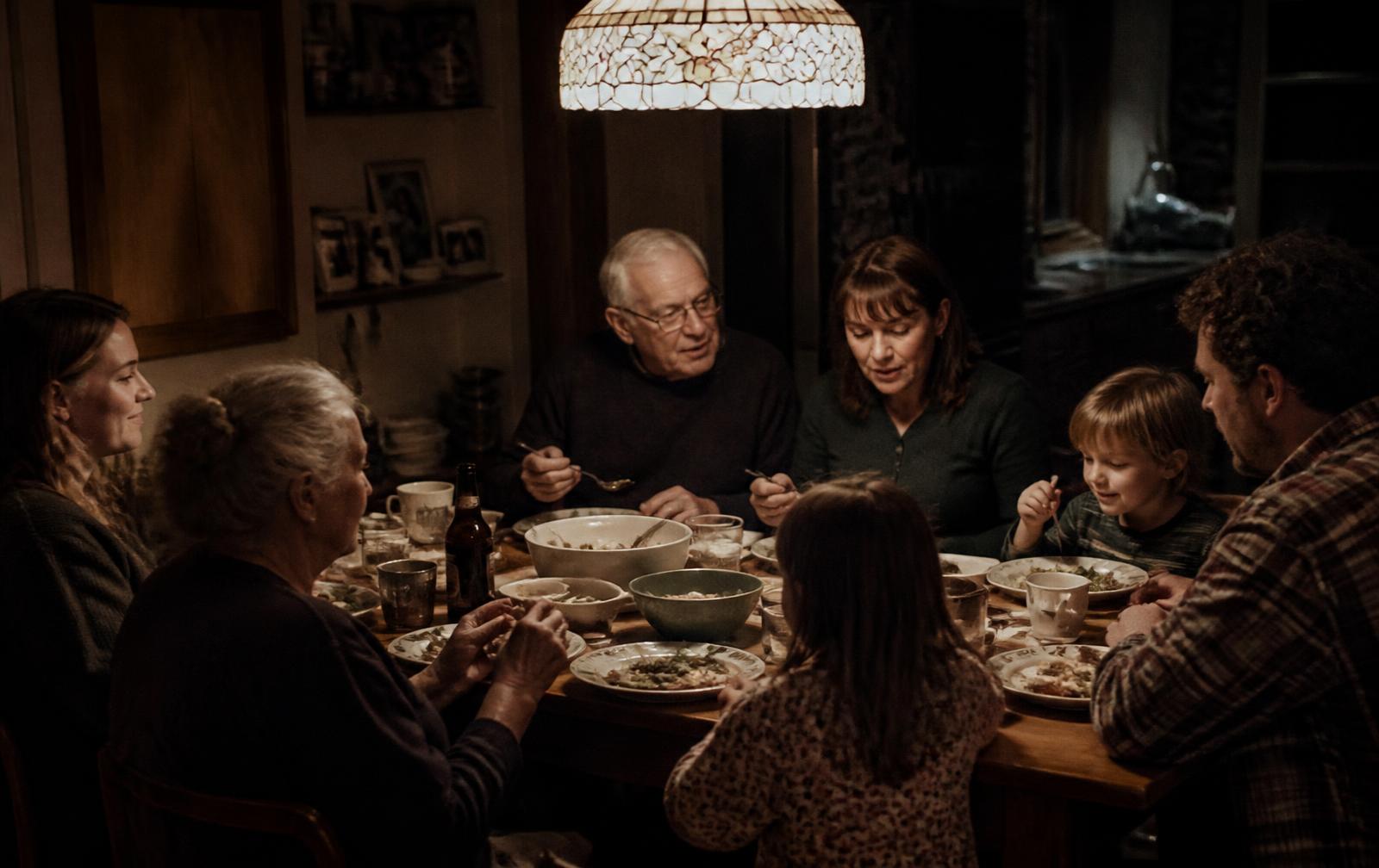 Three generations of a family sharing a meal around a kitchen table under warm lamplight
